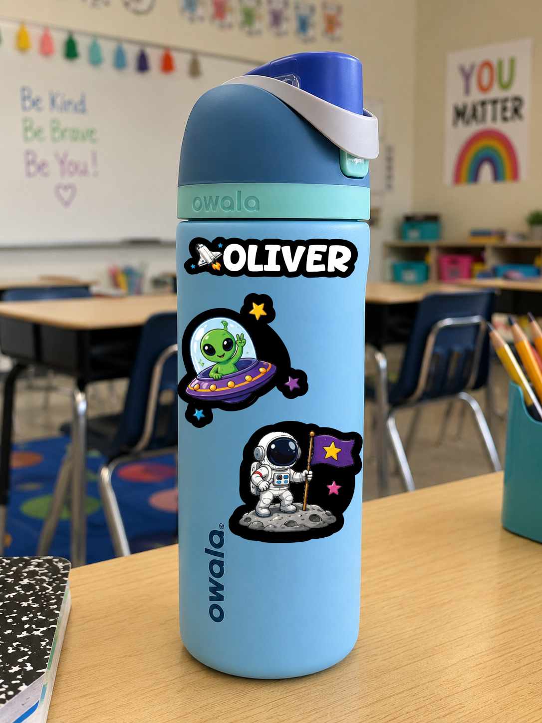 A light blue Owala water bottle with a custom name 'OLIVER' and space-themed stickers, standing on a wooden desk in a classroom.