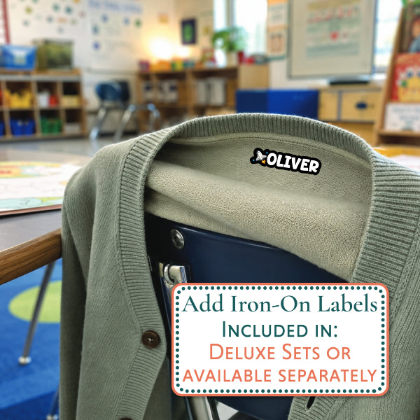An olive green knit cardigan sweater draped over a dark chair in a blurred classroom setting, with an iron-on label sticker reading OLIVER visible on the collar. A text box in the foreground says the labels are included with deluxe sets or available separately.