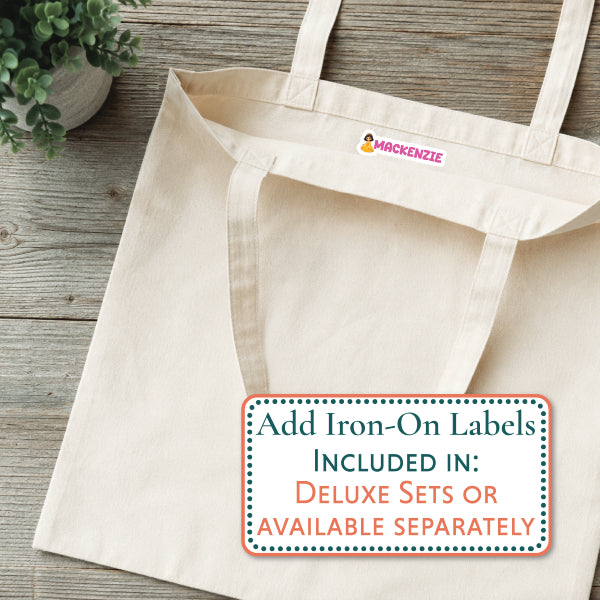 A natural canvas tote bag laid flat on a rustic wood surface, with a small red and yellow NOAH logo printed inside the neck. A white rectangular overlay at the bottom right reads, 'Add Iron-On Labels INCLUDED IN: DELUXE SETS OR AVAILABLE SEPARATELY.