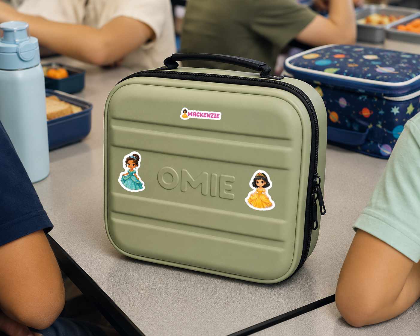 An olive green lunch bag with a black handle and zipper, decorated with stickers and the name 'OMIE' embossed on the front, sitting on a school desk with children around it.