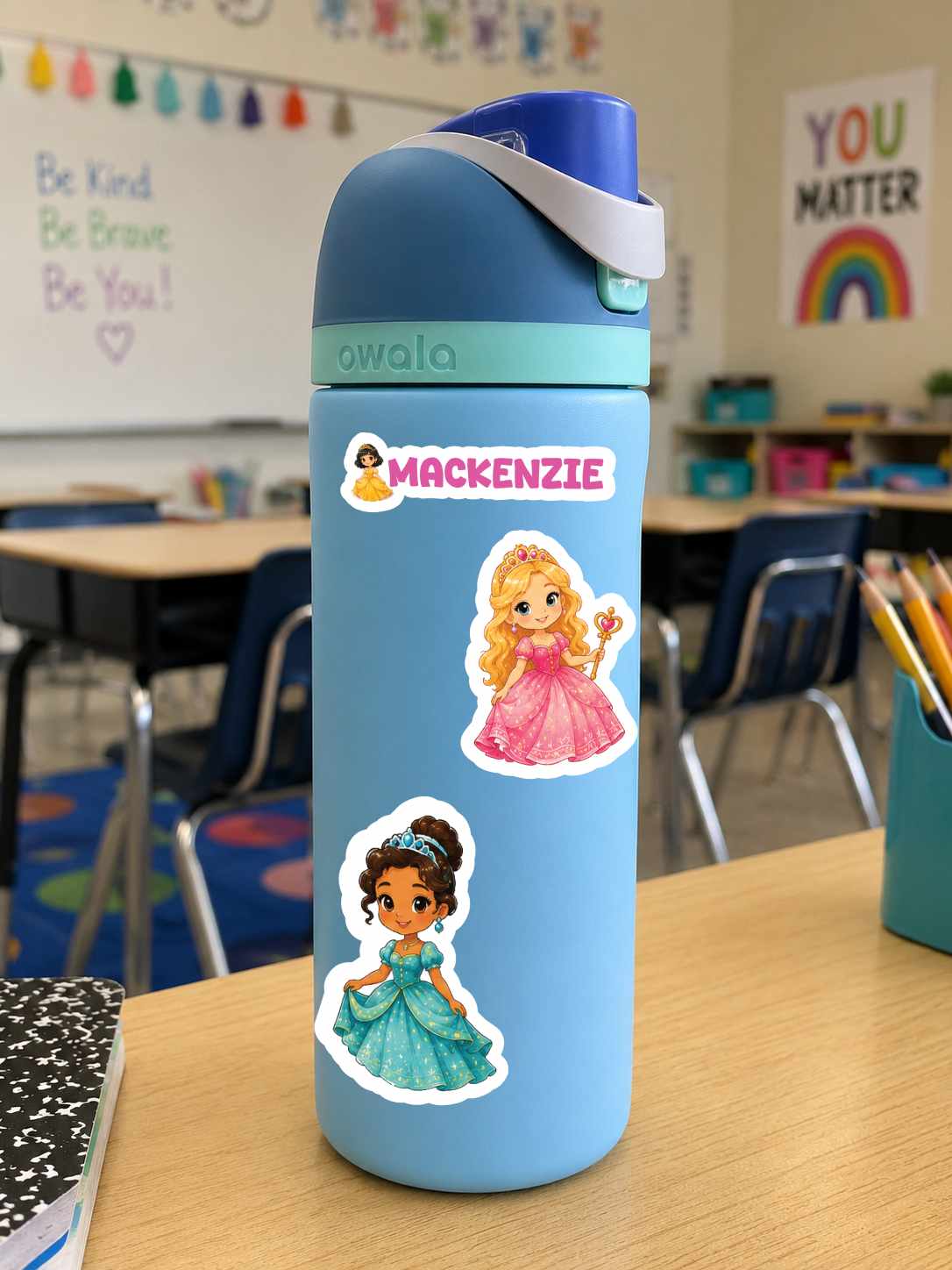 A light blue Owala water bottle with a dark blue lid and a white carrying strap, decorated with two princess stickers (Rapunzel and Tiana) and a custom name sticker that reads 'MACKENZIE'. The bottle is sitting on a wooden desk in a classroom setting.