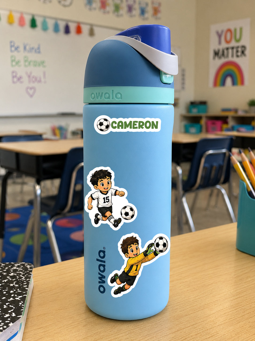A light blue Owala water bottle with a dark blue lid and white strap, decorated with stickers of two soccer players and a 'CAMERON' name label, sitting on a wooden desk in a classroom.