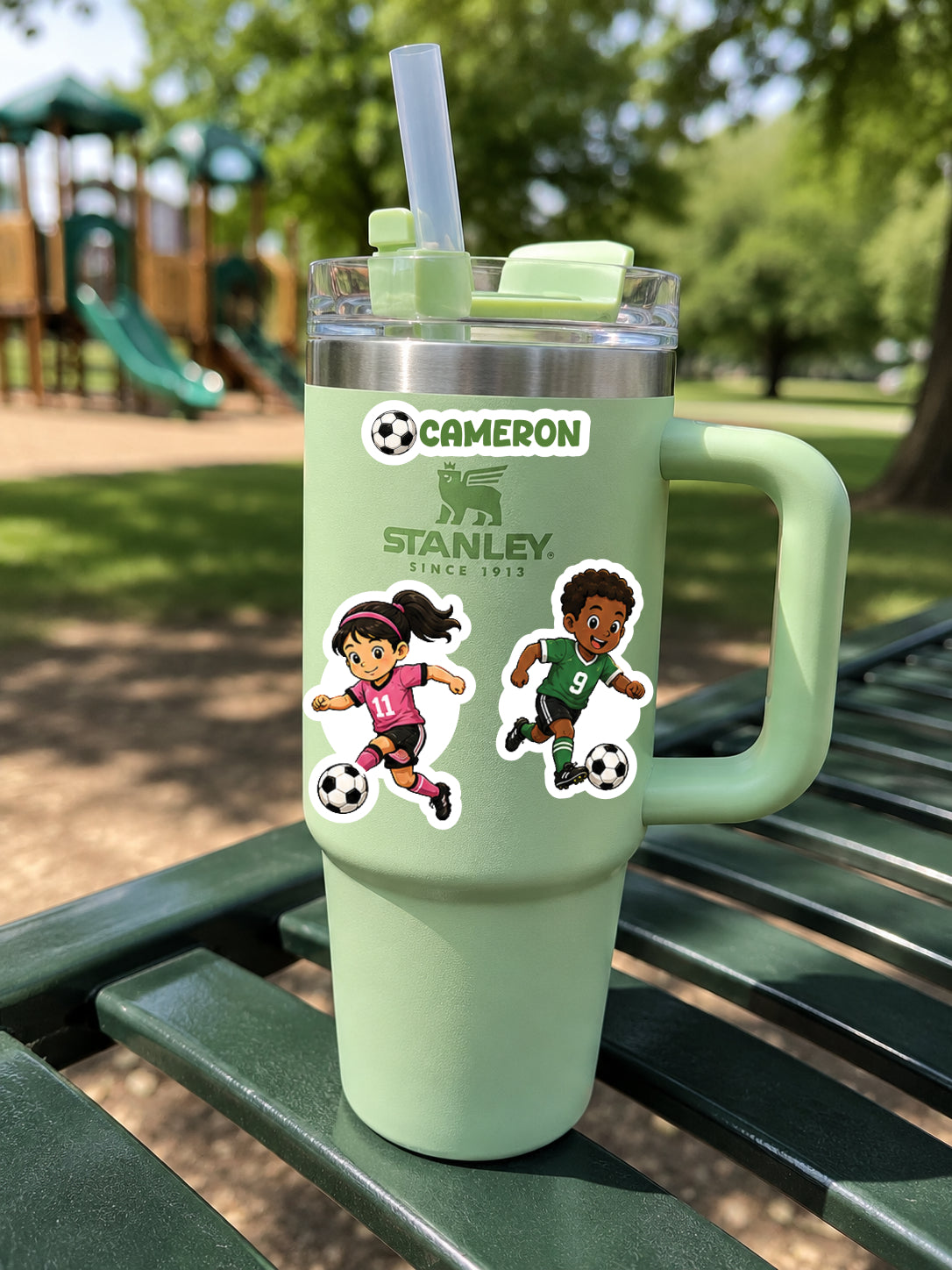 A mint green Stanley tumbler with a clear lid and straw, featuring custom cartoon player stickers and the names 'CAMERON' and 'STANLEY', sitting on a green park bench with a playground in the background.